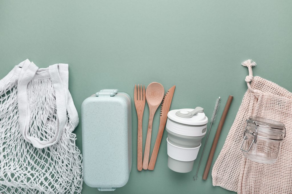 A selection of eco-friendly reusable products arranged on a mint green background. The items include a white mesh tote bag, a light green reusable lunchbox, bamboo utensils (fork, spoon, knife), a collapsible cup with a lid, a reusable straw with a cleaner, and a glass jar in a beige drawstring bag.