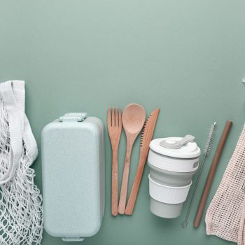 A selection of eco-friendly reusable products arranged on a mint green background. The items include a white mesh tote bag, a light green reusable lunchbox, bamboo utensils (fork, spoon, knife), a collapsible cup with a lid, a reusable straw with a cleaner, and a glass jar in a beige drawstring bag.