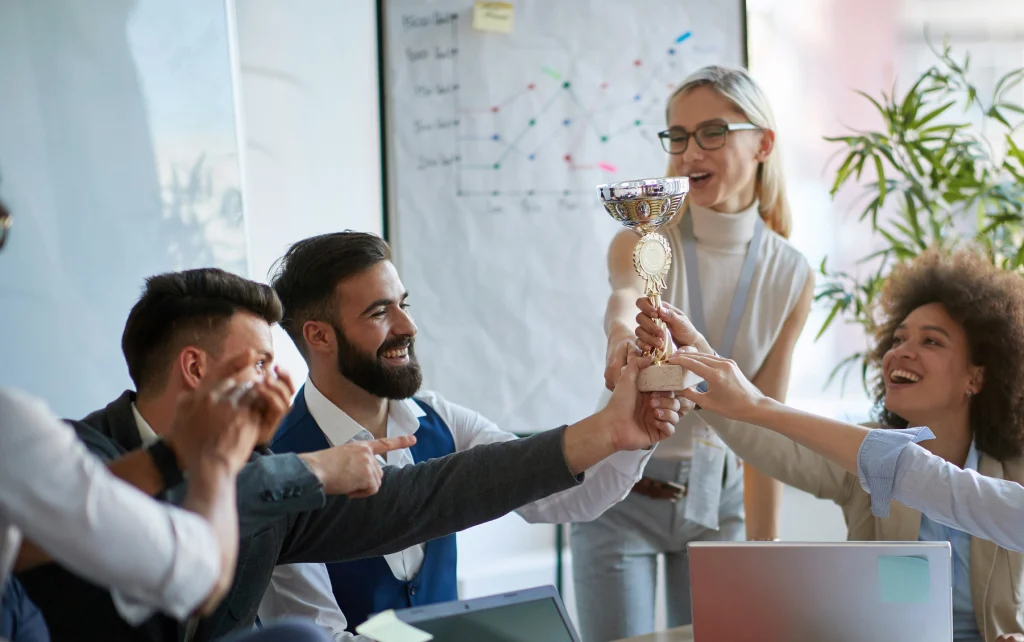 A group of smiling colleagues celebrating together as they hold up a trophy in an office setting, symbolizing teamwork and achievement.