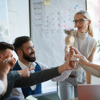 A group of smiling colleagues celebrating together as they hold up a trophy in an office setting, symbolizing teamwork and achievement.