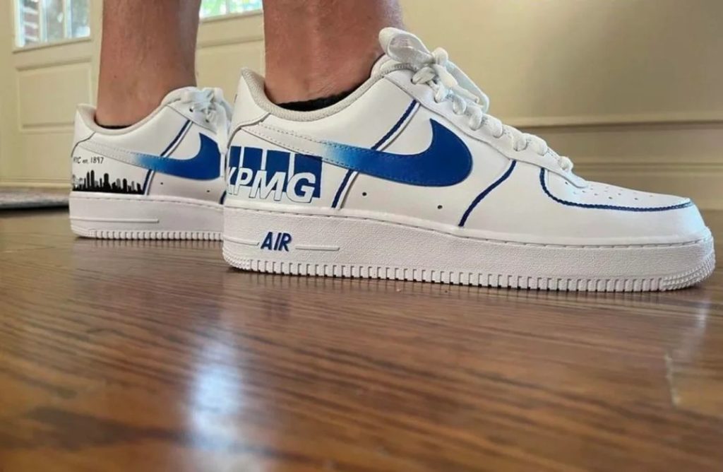 White Nike Air Force 1 sneakers customised with the KPMG logo and blue swoosh design.