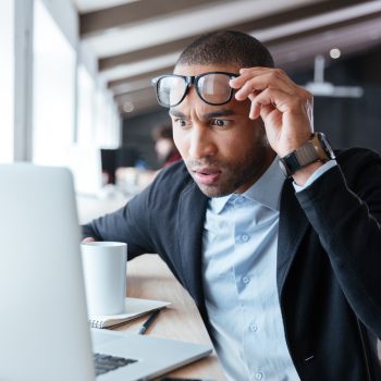 A surprised man lifting his glasses while looking at a laptop screen with a cup of coffee beside him.