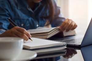 A person writing in a notebook while reading a book, with a laptop in the background, suggesting focused study or work.