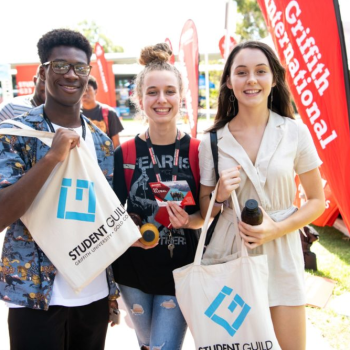 Three students smiling and holding Student Guild tote bags at an outdoor university event, surrounded by Griffith University banners.