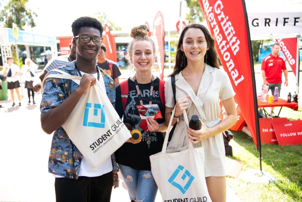 Three students smiling and holding Student Guild tote bags at an outdoor university event, surrounded by Griffith University banners.